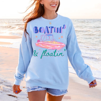 Woman walking on the beach wearing a light blue Boatin’ & Floatin’ sweatshirt featuring retro pink boat artwork and coastal summer style.