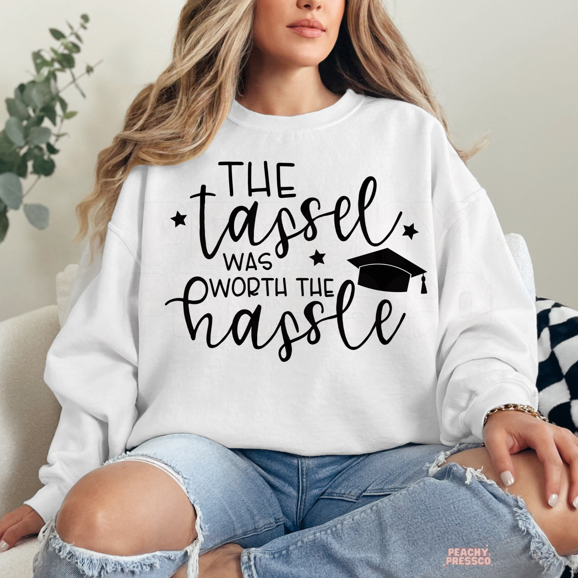 Woman wearing white sweatshirt featuring The Tassel Was Worth The Hassle graduation quote with graduation cap graphic.