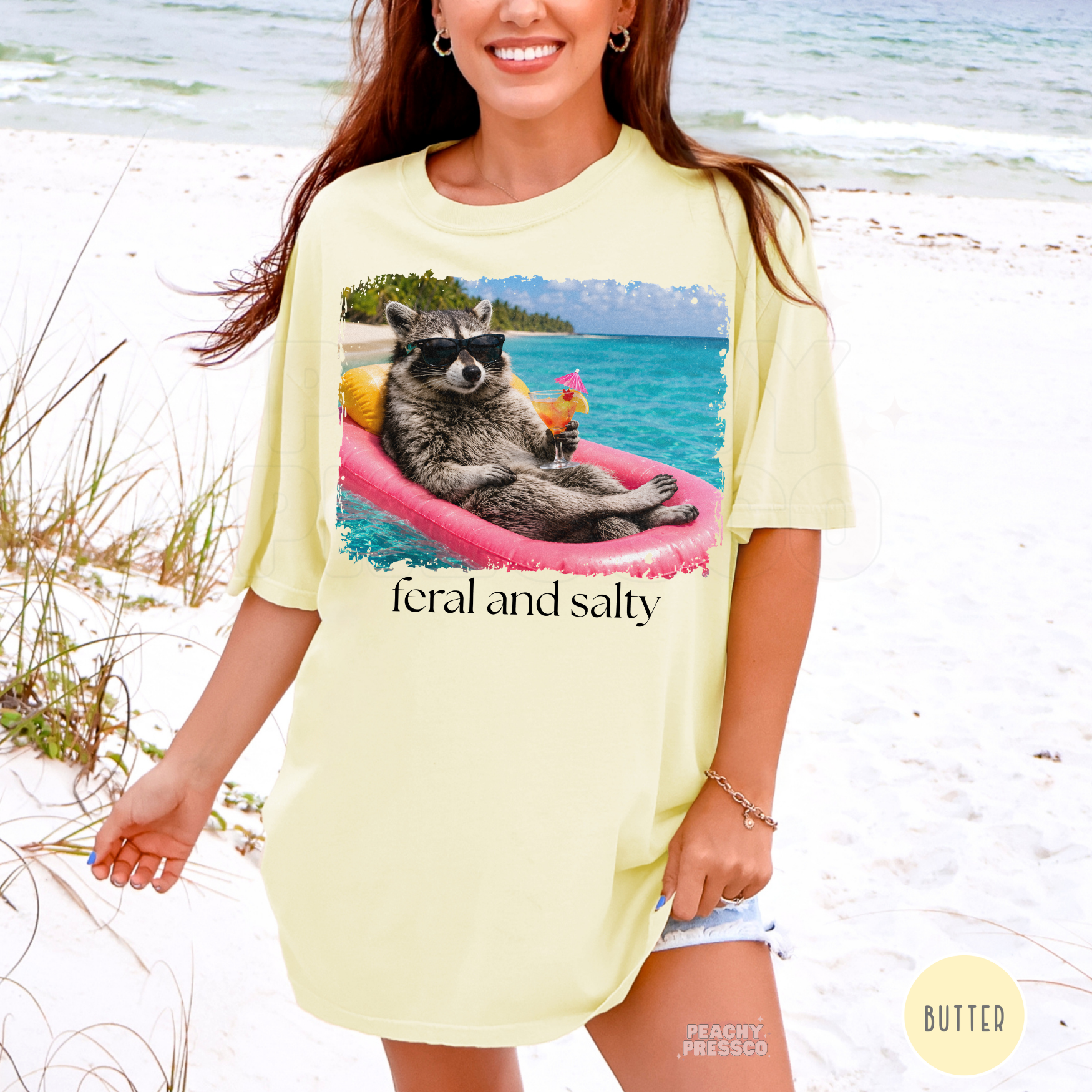 Woman wearing a butter yellow oversized beach t-shirt featuring a floating raccoon with tropical drink and “Feral and Salty” summer graphic.