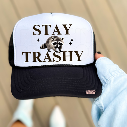 White and black trucker hat featuring raccoon illustration and “Stay Trashy” text.
