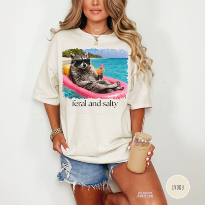 Woman wearing an ivory oversized graphic t-shirt featuring a raccoon floating on a pink raft with ocean background and “Feral and Salty” design.