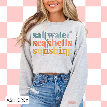 Woman wearing an ash grey sweatshirt featuring retro “saltwater seashells sunshine” text in pastel coastal-inspired colors.