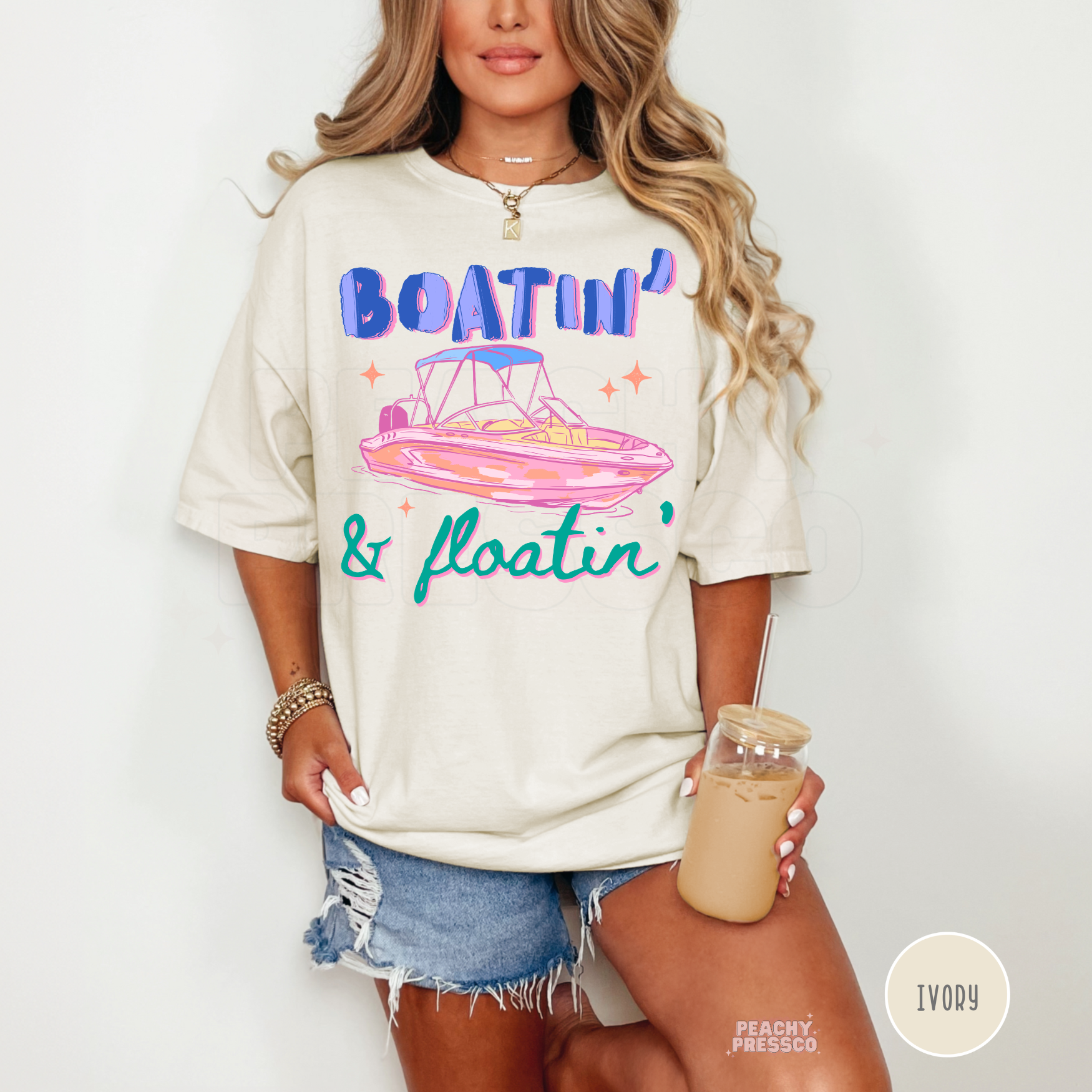 Woman wearing an ivory oversized graphic t-shirt featuring Boatin’ & Floatin’ retro pink boat artwork styled with denim shorts and iced coffee.