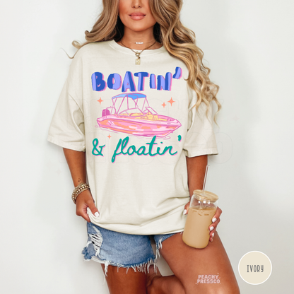 Woman wearing an ivory oversized graphic t-shirt featuring Boatin’ & Floatin’ retro pink boat artwork styled with denim shorts and iced coffee.