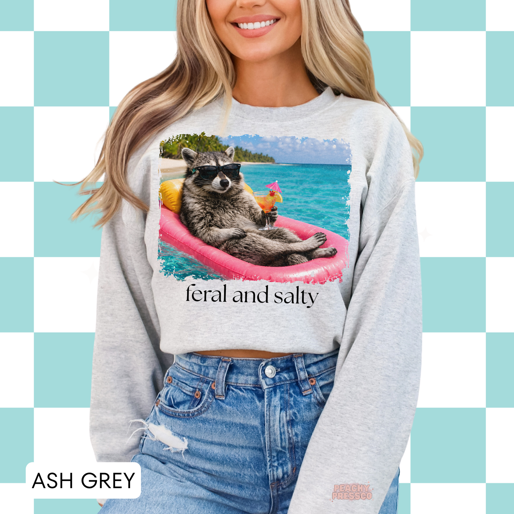 Woman wearing an ash grey sweatshirt featuring a tropical raccoon float design with sunglasses and “Feral and Salty” graphic.