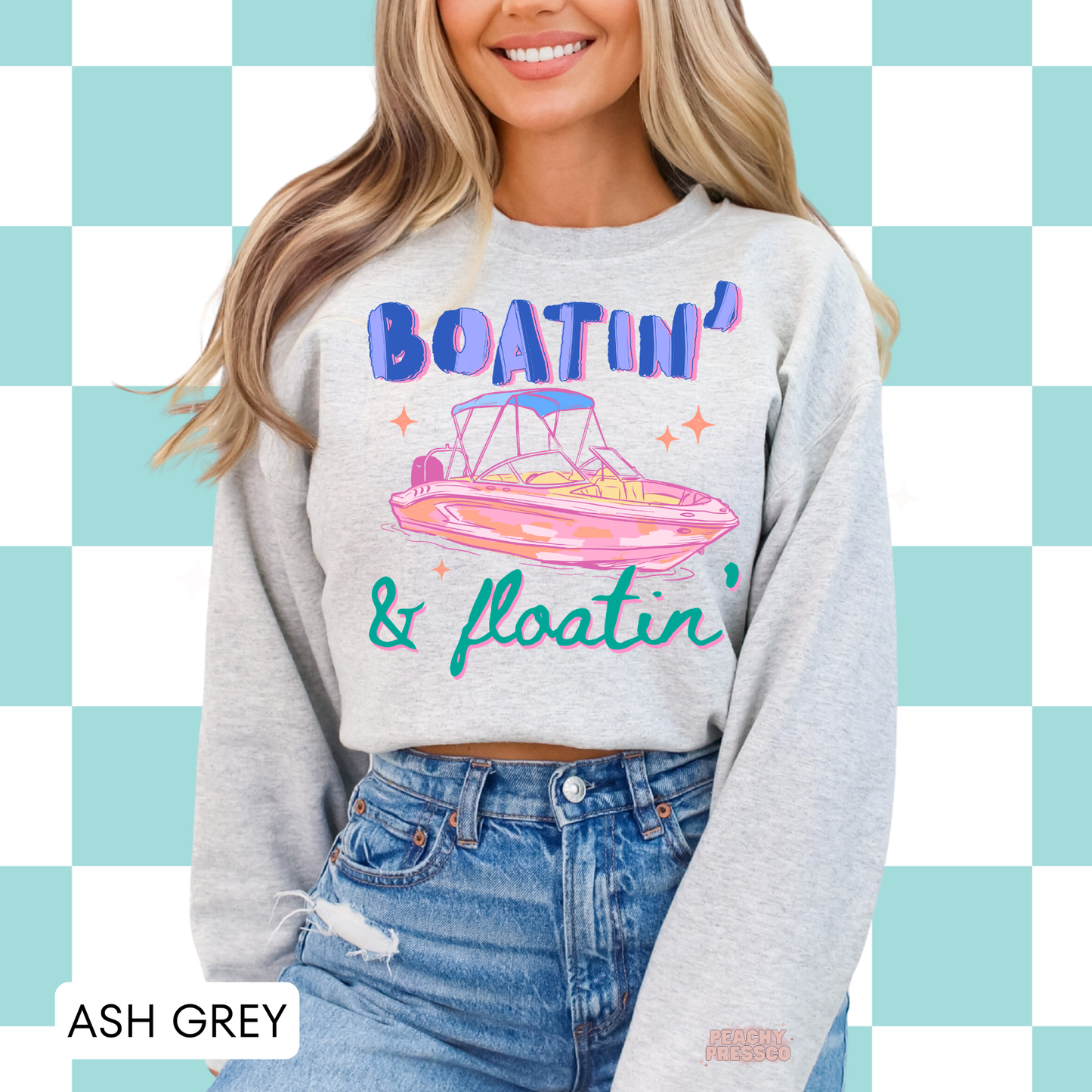 Woman wearing an ash grey crewneck sweatshirt featuring retro “Boatin’ & Floatin’” graphic with pink boat illustration and colorful lettering.