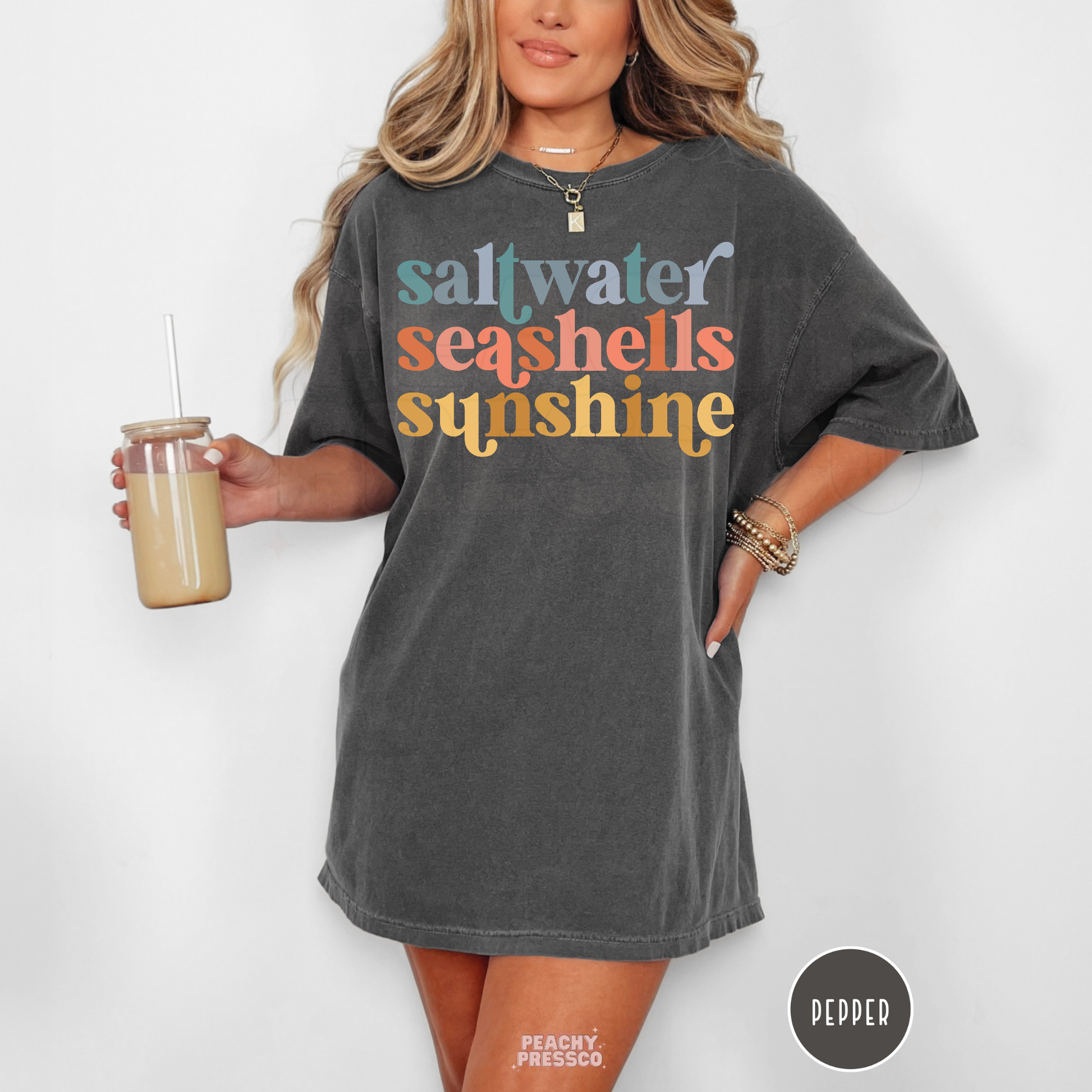 Woman wearing a pepper grey oversized t-shirt featuring retro “saltwater seashells sunshine” typography in soft pastel summer colors.