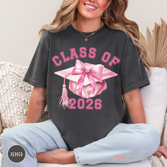 Trendy Class of 2026 graduation t-shirt with pink bow cap illustration, varsity lettering, and coquette-inspired graduate apparel.
