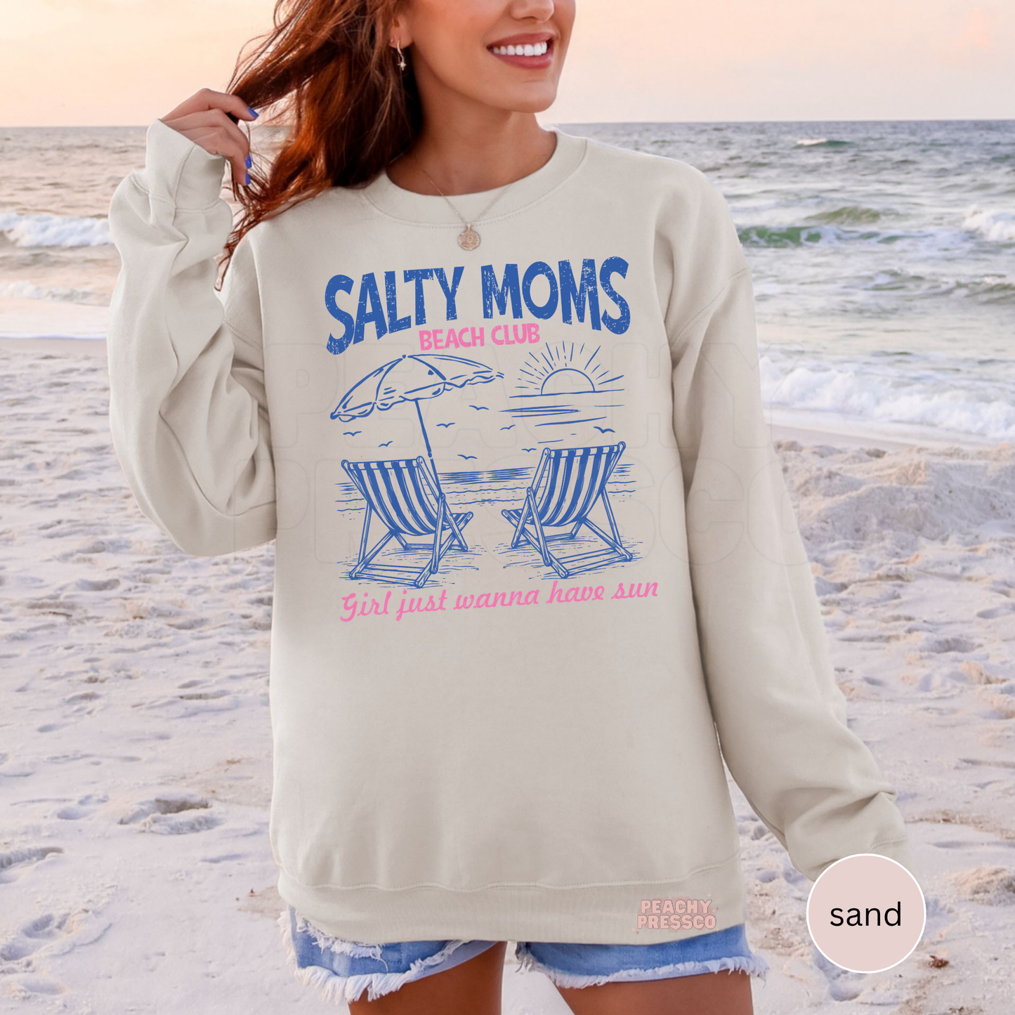 Woman wearing a sand colored crewneck sweatshirt with retro beach club graphic featuring lounge chairs, ocean sunset, and pink lettering.