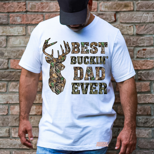 Man wearing white t-shirt with camo deer head graphic and “Best Buckin’ Dad Ever” text standing in front of brick wall.
