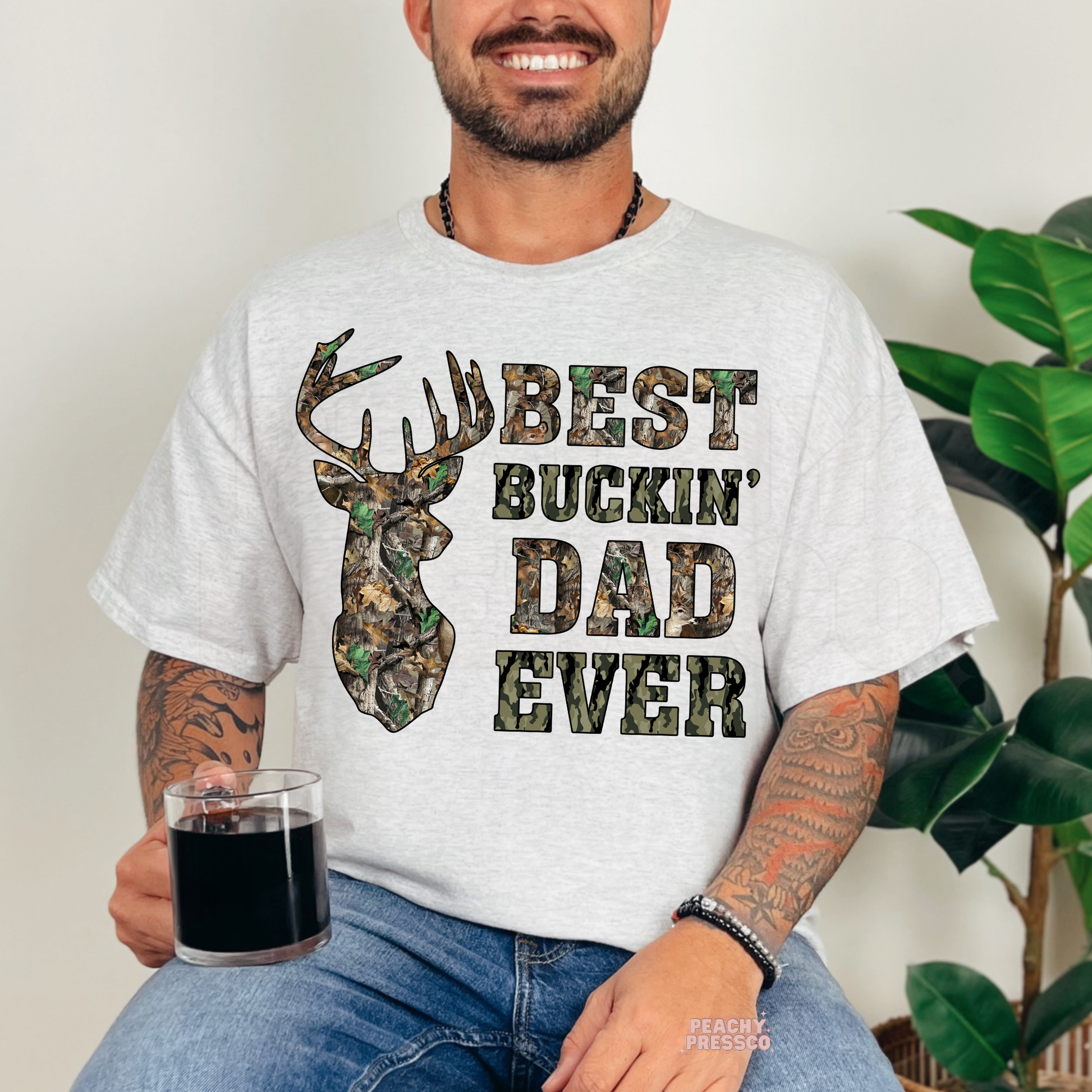 Man sitting indoors wearing ash grey t-shirt featuring camo deer silhouette and “Best Buckin’ Dad Ever” hunting-inspired graphic.