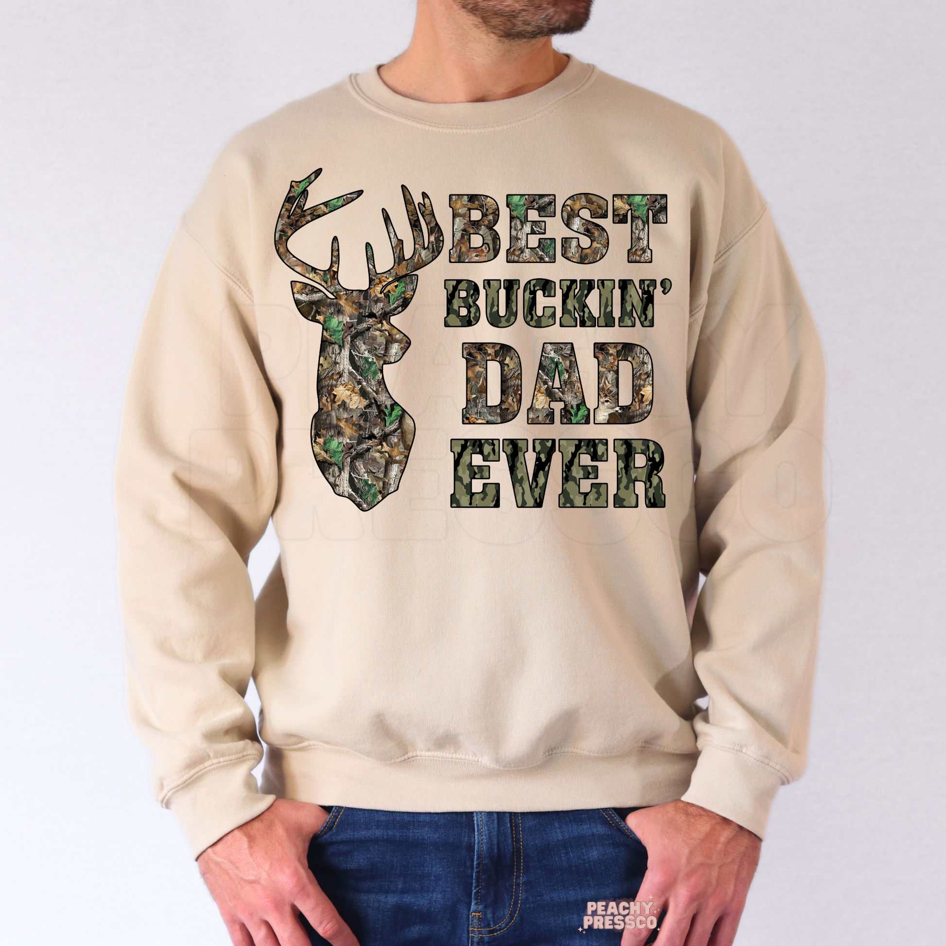 Man wearing beige sweatshirt with rustic camouflage “Best Buckin’ Dad Ever” deer hunting graphic design.