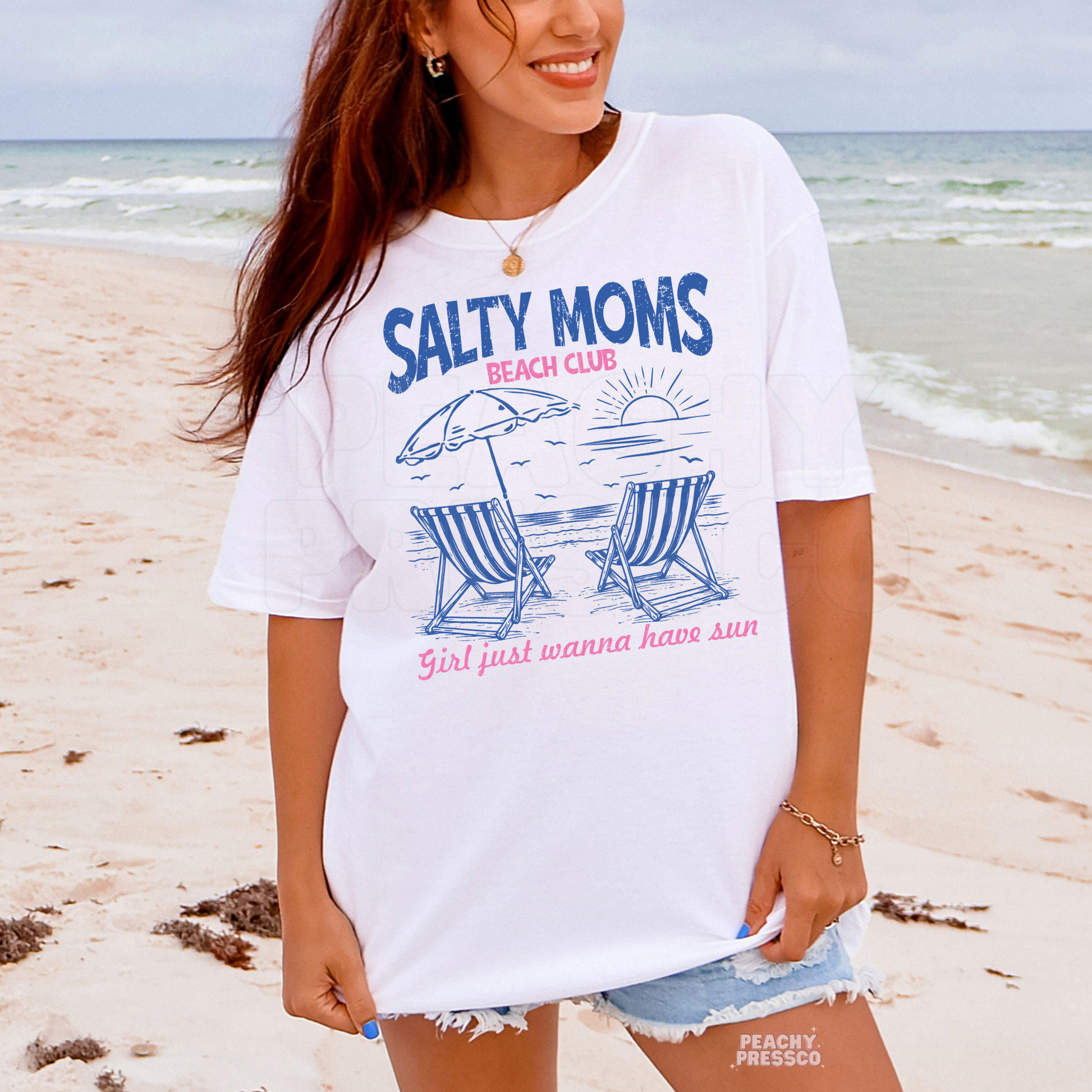 Woman wearing a white oversized beach t-shirt featuring “Salty Moms Beach Club” graphic with coastal chairs and sunset artwork.