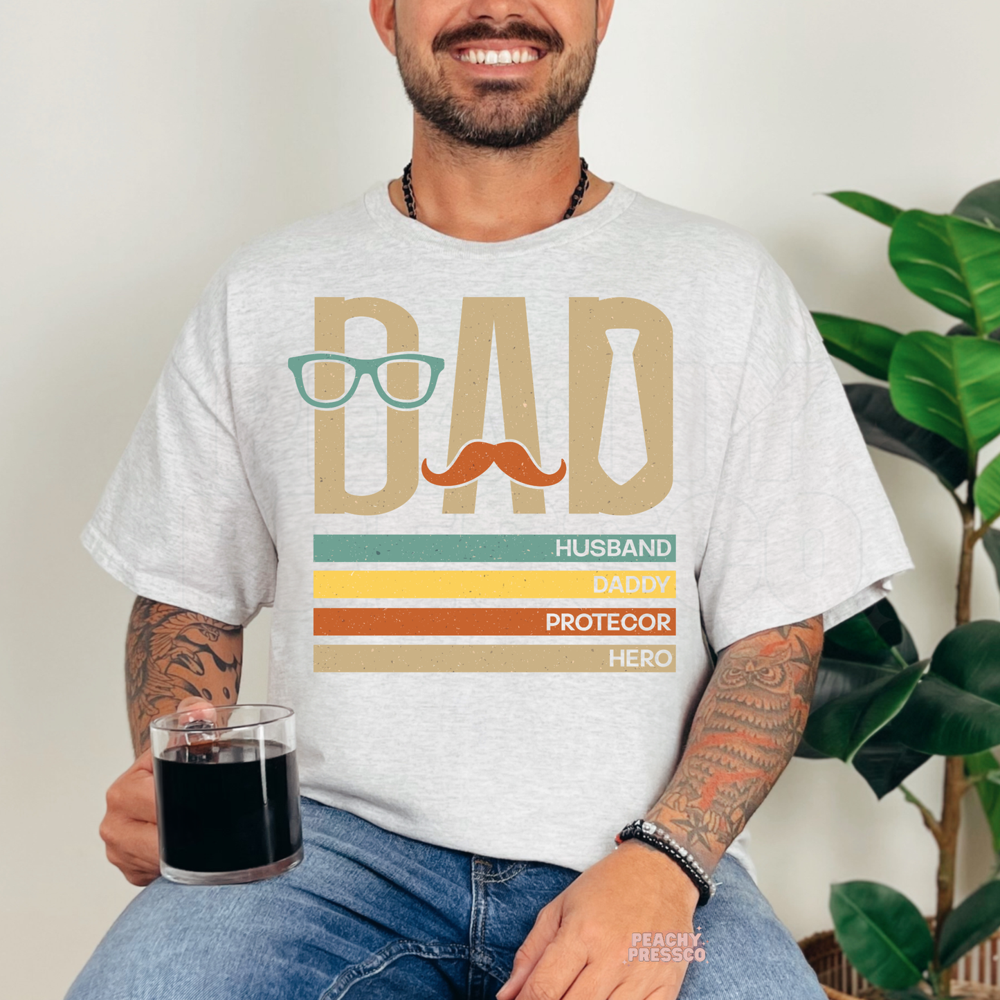 Man wearing ash grey t-shirt with retro dad graphic featuring glasses, mustache, tie, and colorful “Husband Daddy Protector Hero” text.