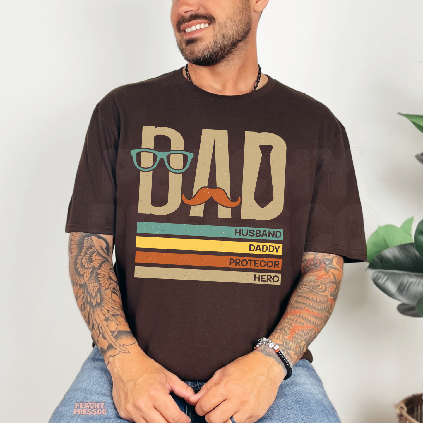 Man sitting indoors wearing dark brown t-shirt with vintage-style “Dad Husband Daddy Protector Hero” Father’s Day graphic.