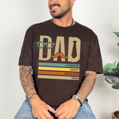 Man sitting indoors wearing dark brown t-shirt with vintage-style “Dad Husband Daddy Protector Hero” Father’s Day graphic.