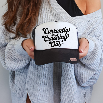 Black and white trucker hat featuring “Currently Crashing Out” script text.
