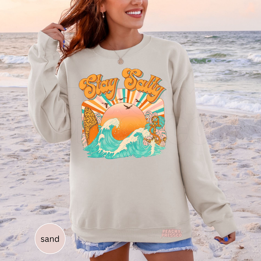 Woman wearing a sand colored crewneck sweatshirt featuring vintage style “Stay Salty” beach artwork with waves, sunset, and tropical details.