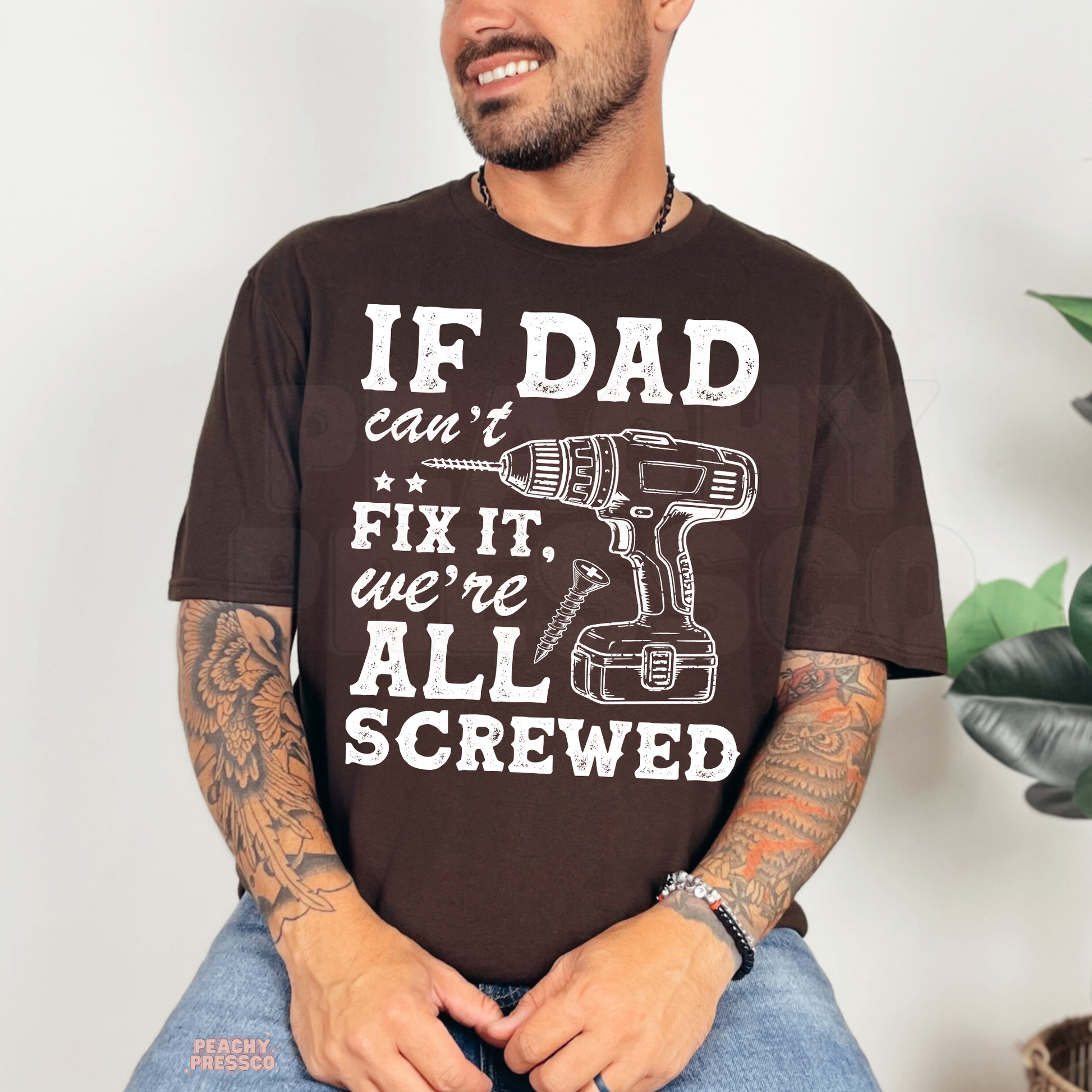 Smiling man modeling dark brown t-shirt with funny “If Dad Can’t Fix It” design and power drill graphic.