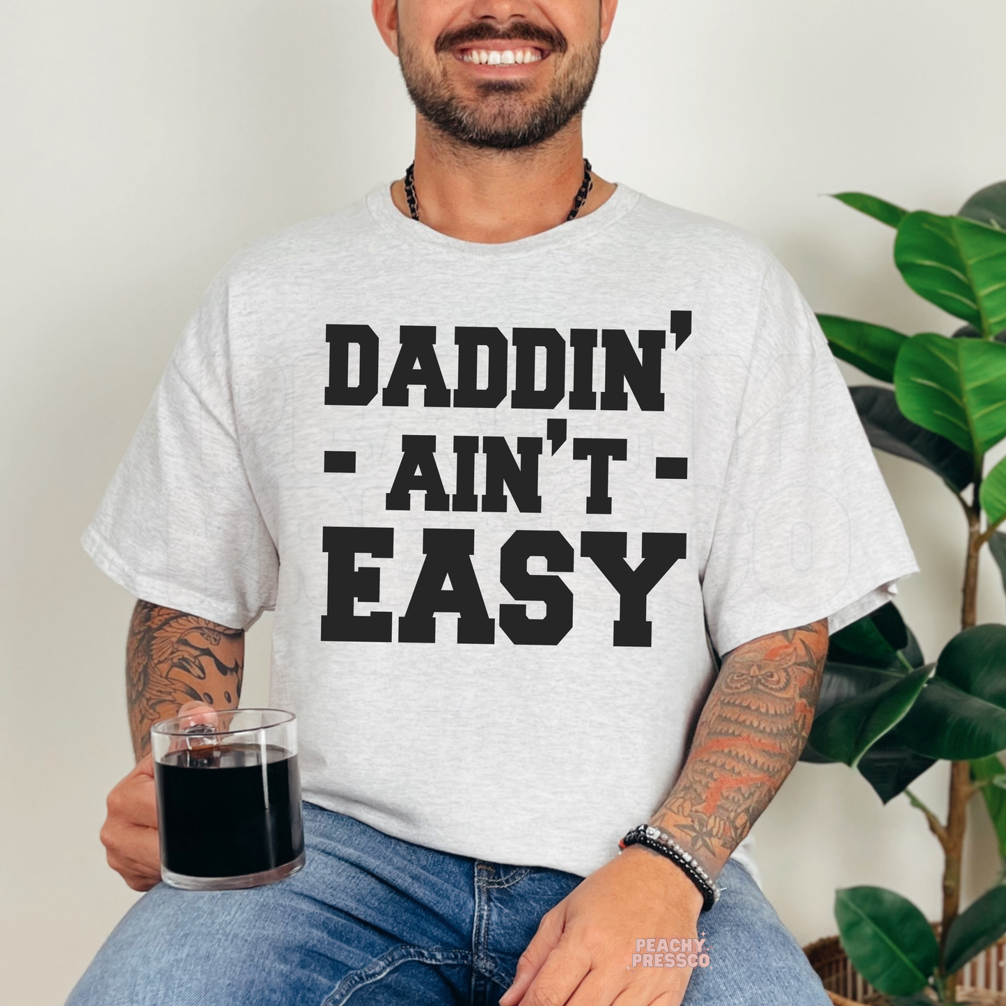 Man wearing ash grey t-shirt with bold black “Daddin’ Ain’t Easy” slogan graphic and holding a glass mug.