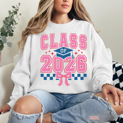 Woman wearing white sweatshirt featuring pink Class of 2026 graduation design with bow, checker accents and graduation cap graphic.