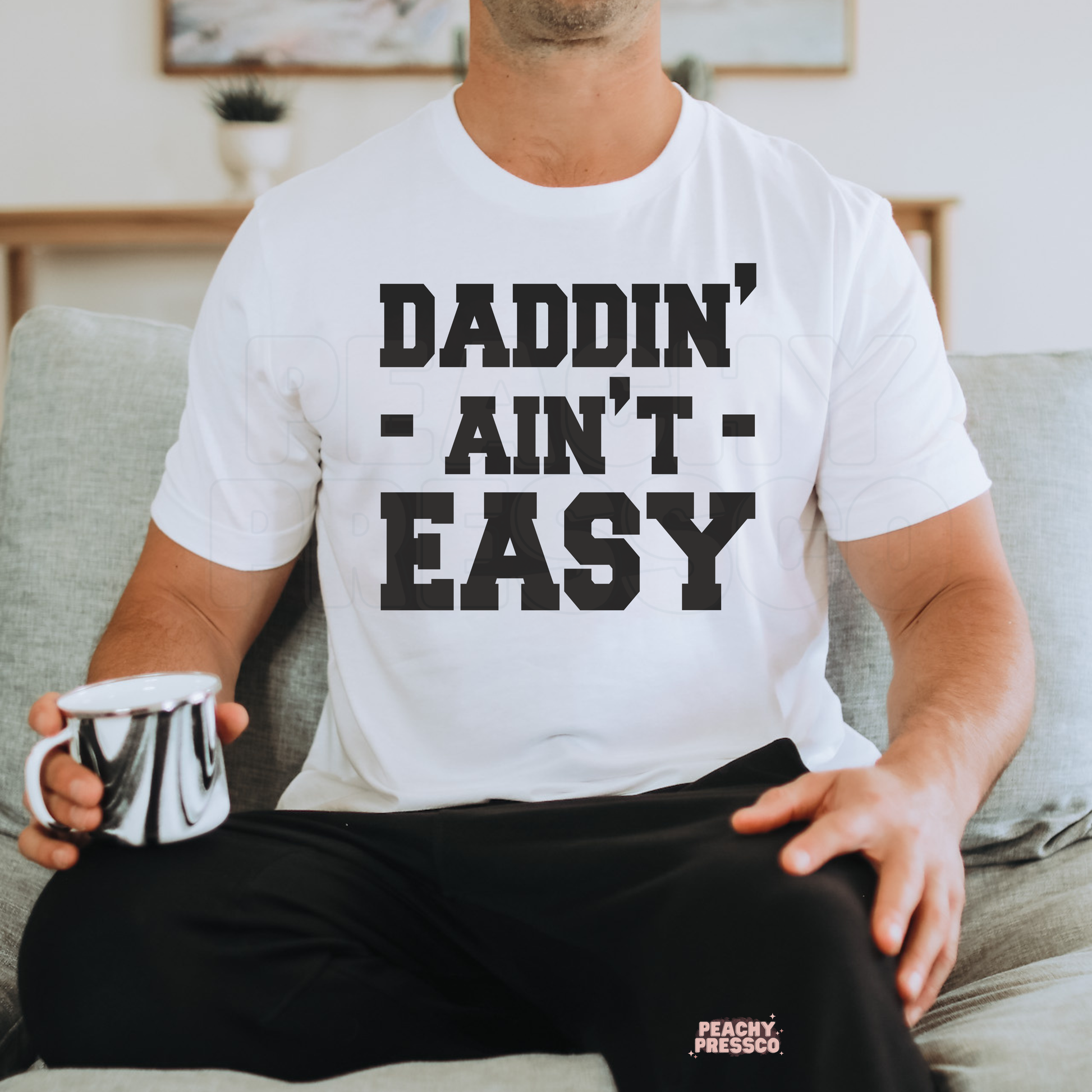 Man sitting on couch wearing white t-shirt with “Daddin’ Ain’t Easy” bold black typography graphic.
