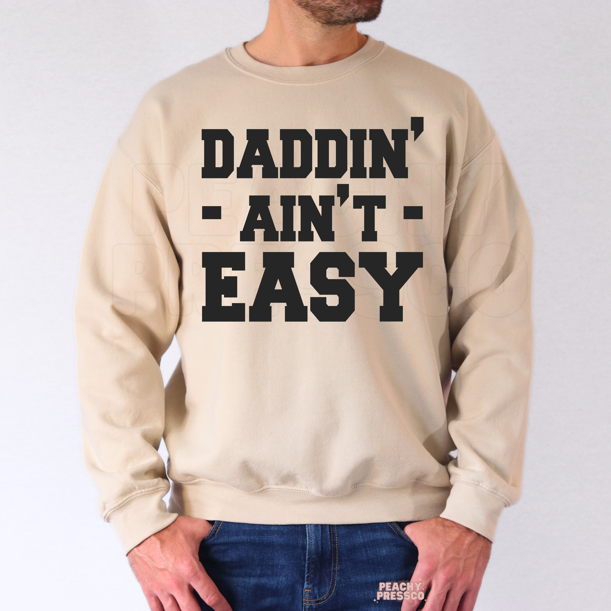 Man wearing sand-colored crewneck sweatshirt with oversized black “Daddin’ Ain’t Easy” Father’s Day graphic.