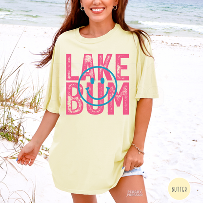 Woman wearing oversized butter yellow Lake Bum graphic t-shirt on the beach with pink distressed lettering and blue smiley face.