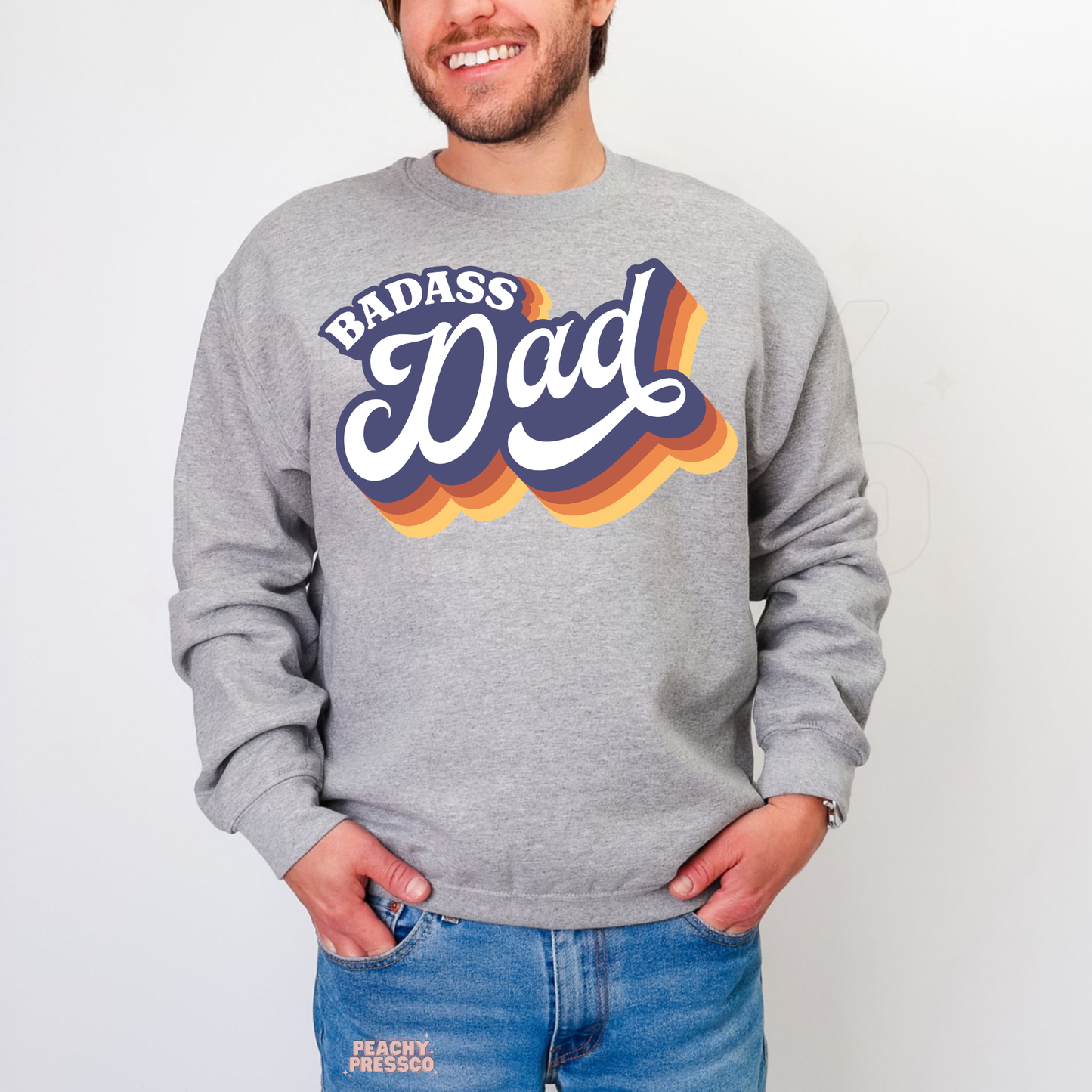 Man wearing grey crewneck sweatshirt with retro “Badass Dad” graphic in layered sunset-inspired colors.