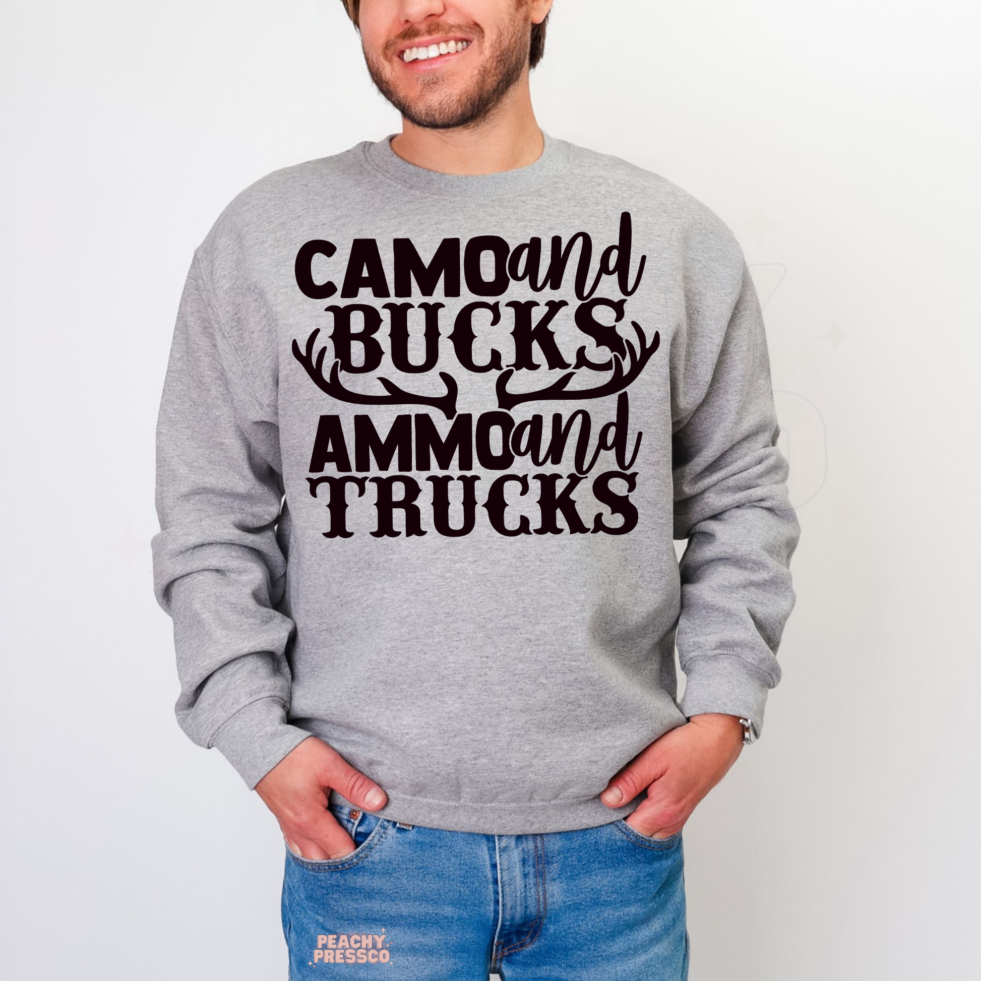 Man wearing grey crewneck sweatshirt with bold “Camo and Bucks Ammo and Trucks” hunting quote graphic.
