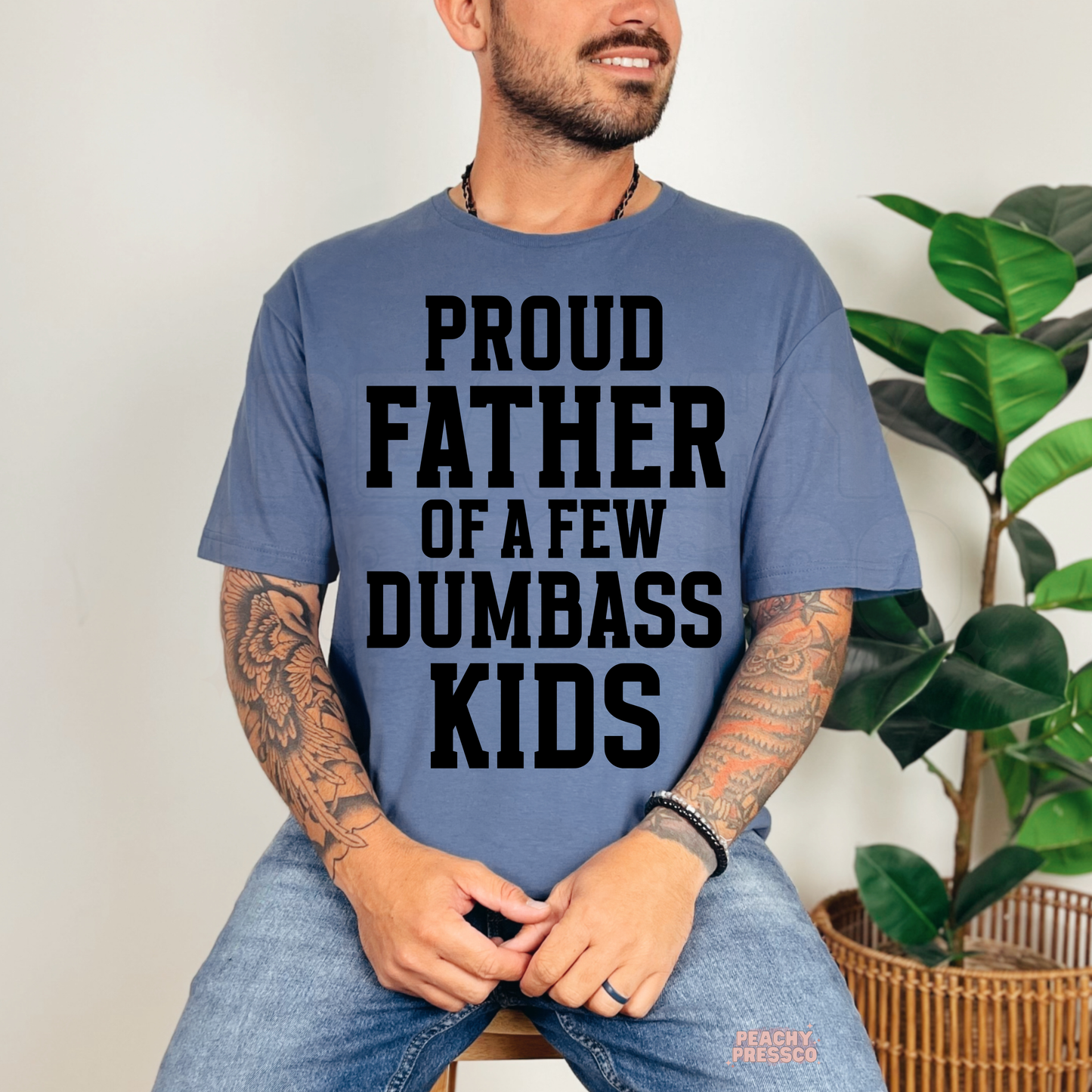 Smiling man wearing blue t-shirt with bold black “Proud Father of a Few Dumbass Kids” graphic typography design.