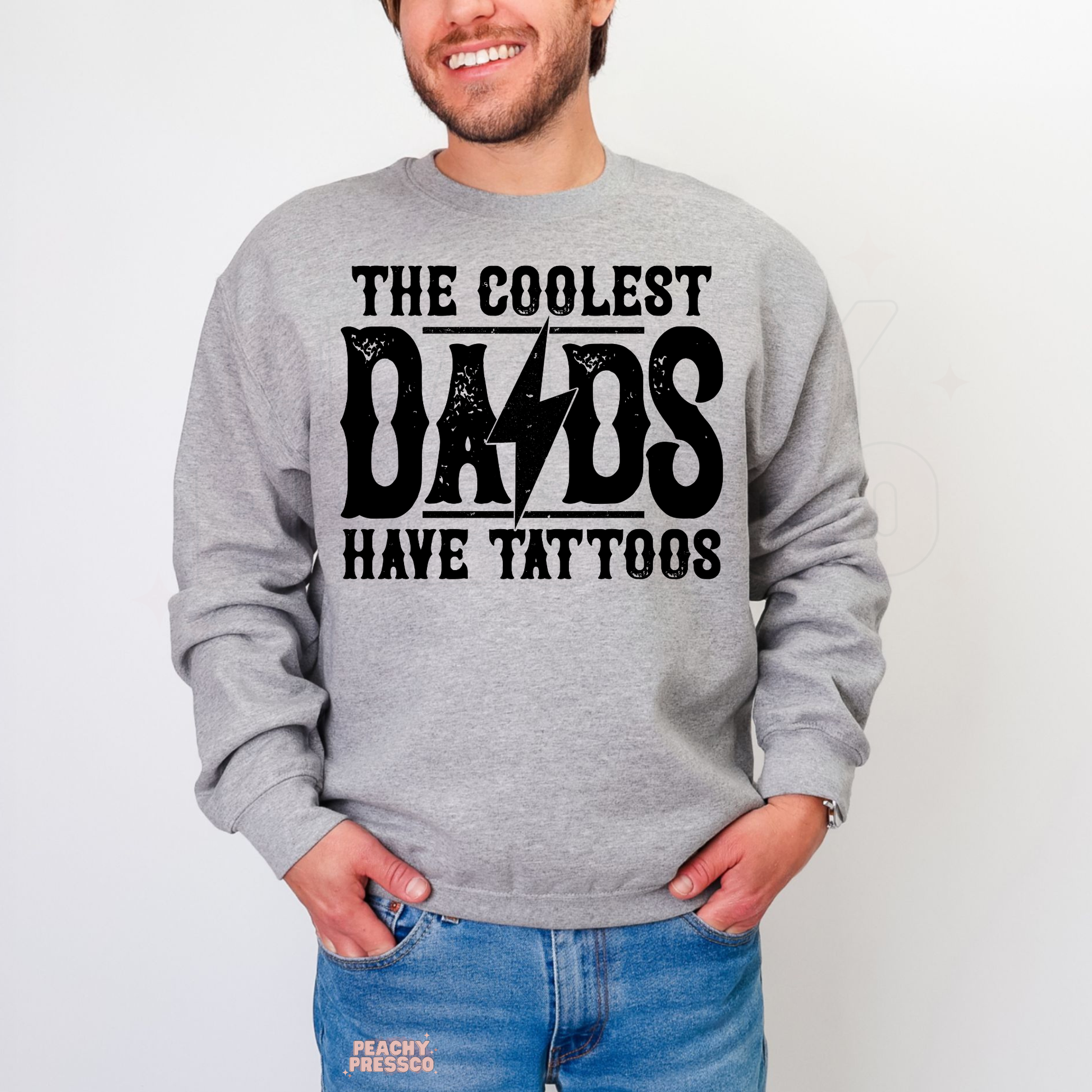 Man modeling grey crewneck sweatshirt with bold “The Coolest Dads Have Tattoos” retro-style graphic.