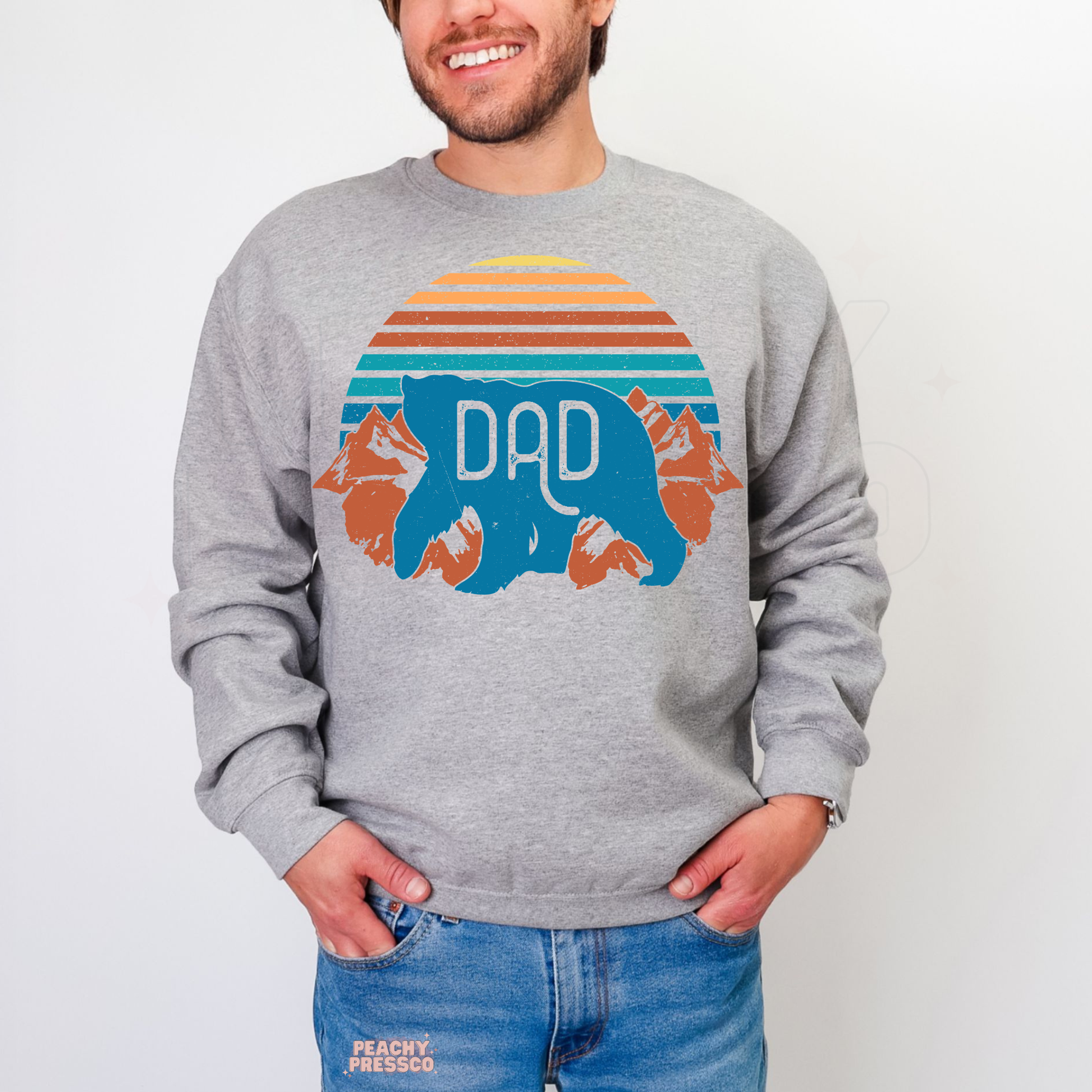 Man wearing grey crewneck sweatshirt featuring retro mountain sunset and bear “Dad” graphic design.