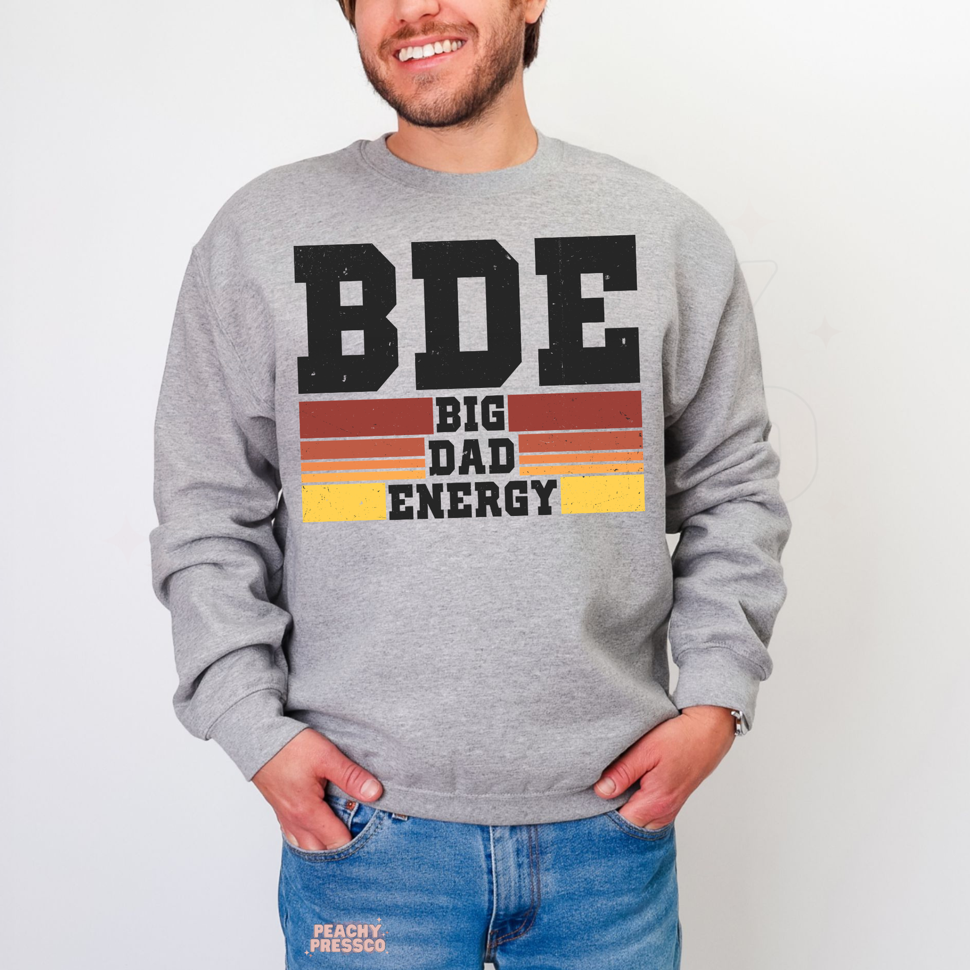 Man wearing grey crewneck sweatshirt with retro “BDE Big Dad Energy” graphic in vintage sunset stripe design.
