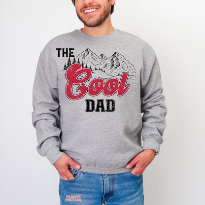 Man modeling grey crewneck sweatshirt with vintage-inspired “The Good Dad” mountain graphic design.