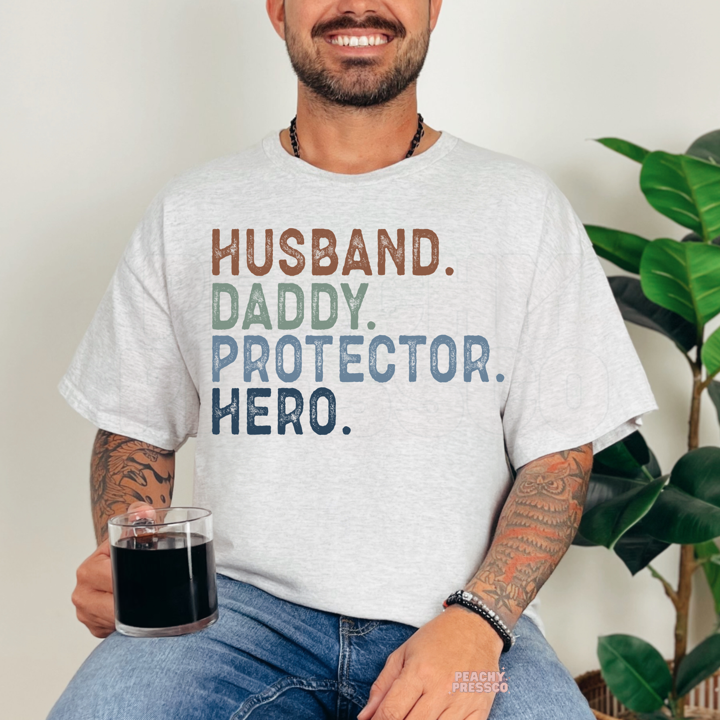 Smiling man wearing ash grey t-shirt with colorful “Husband. Daddy. Protector. Hero.” typography graphic design.