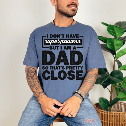 Man modeling blue t-shirt with humorous “Dad Superpowers” graphic design in bold distressed lettering.