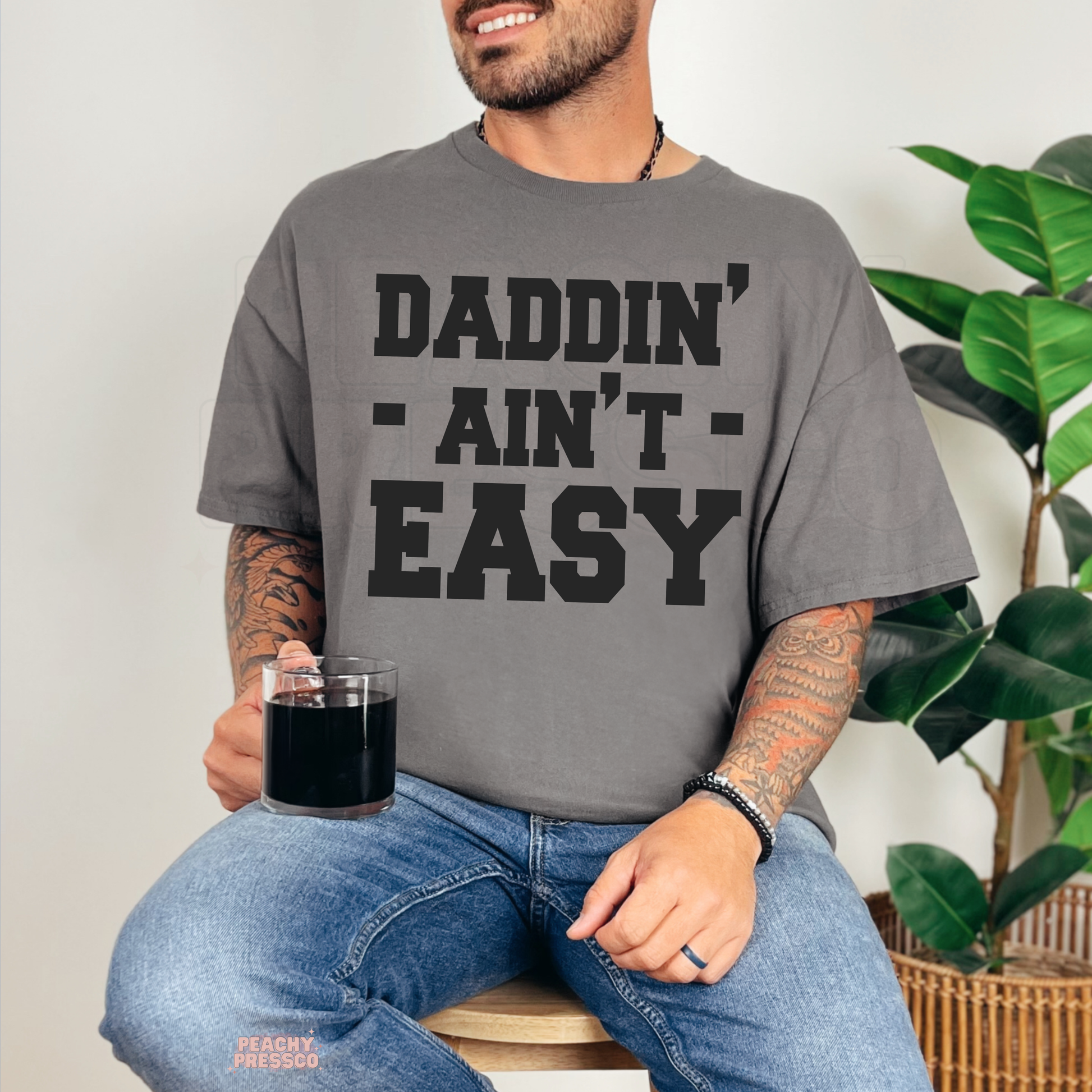 Man sitting indoors wearing charcoal grey t-shirt featuring bold “Daddin’ Ain’t Easy” dad humor graphic.