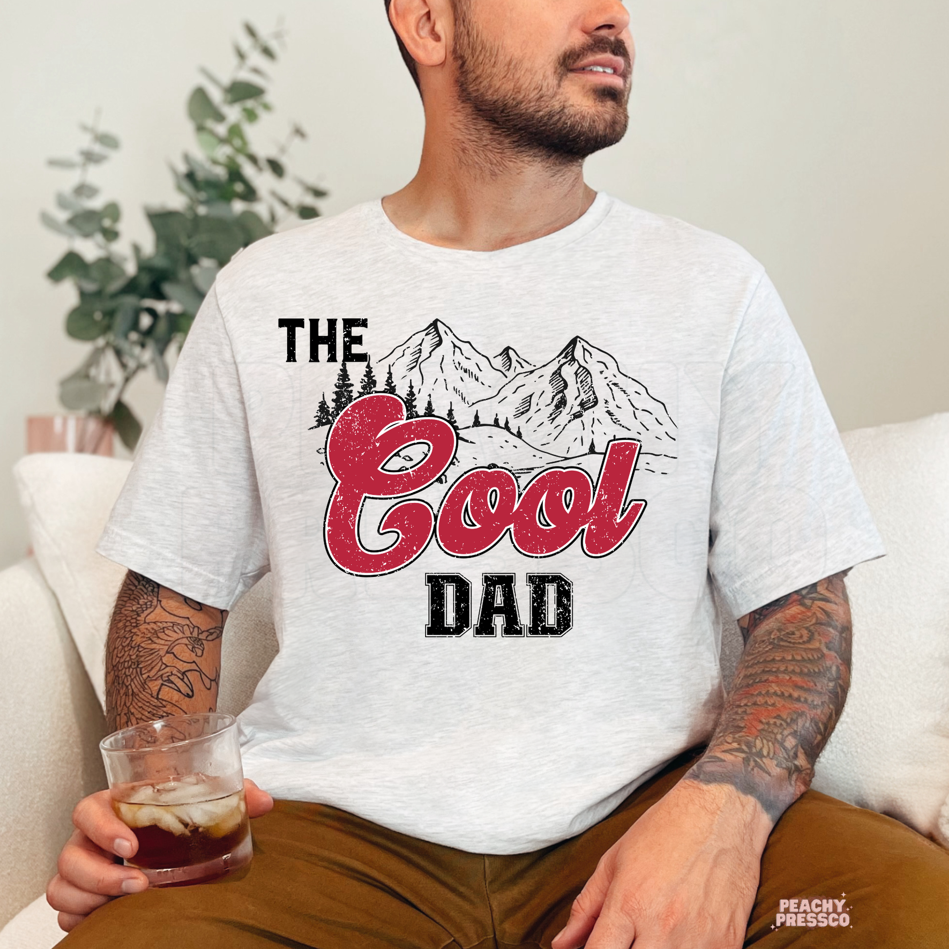 Man sitting indoors wearing ash grey t-shirt with retro “The Good Dad” graphic and mountain artwork.