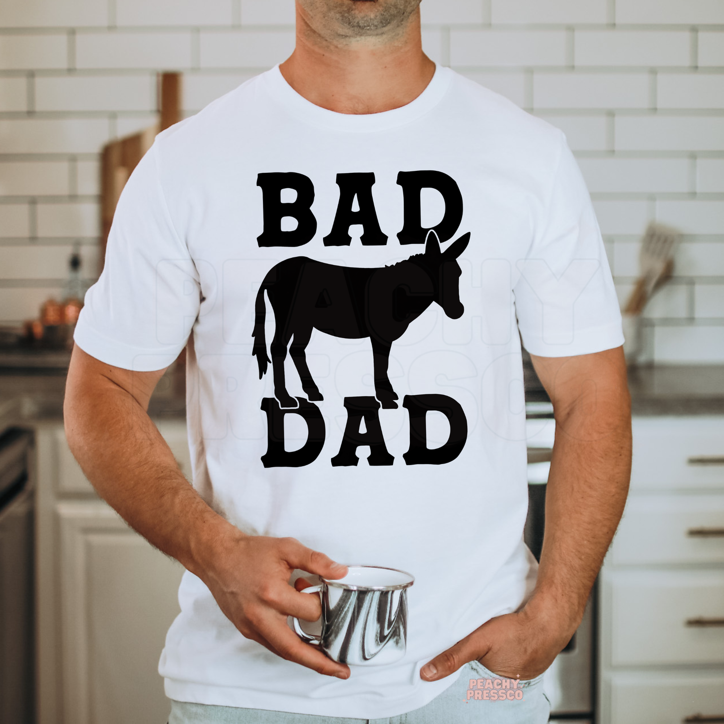 Man wearing white t-shirt with bold black “Bad Ass Dad” graphic and donkey silhouette while holding a metal mug.