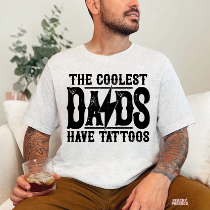 Man sitting indoors wearing ash grey t-shirt with “The Coolest Dads Have Tattoos” statement graphic.