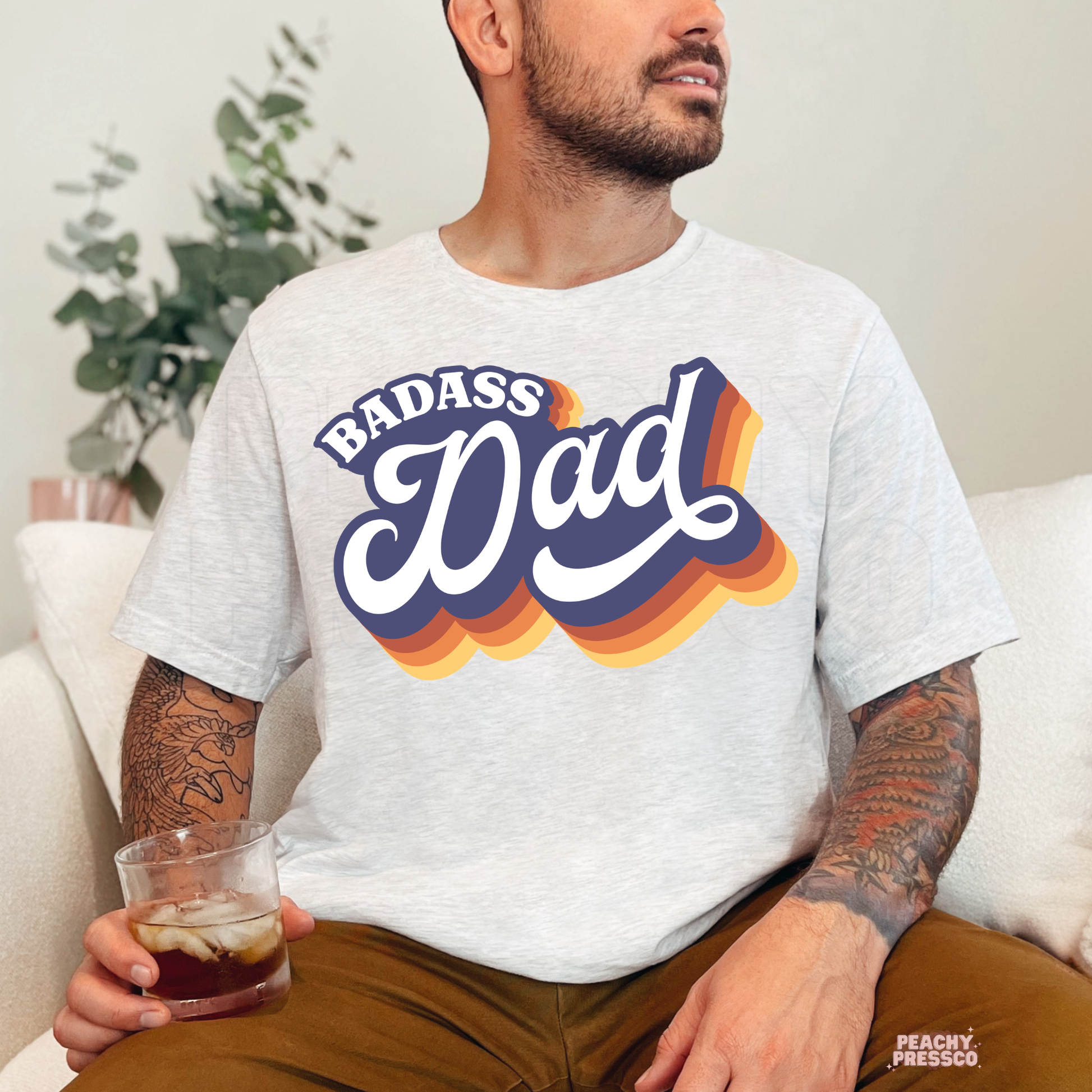 Man sitting indoors wearing ash grey t-shirt with bold retro “Badass Dad” graphic and vintage style lettering.