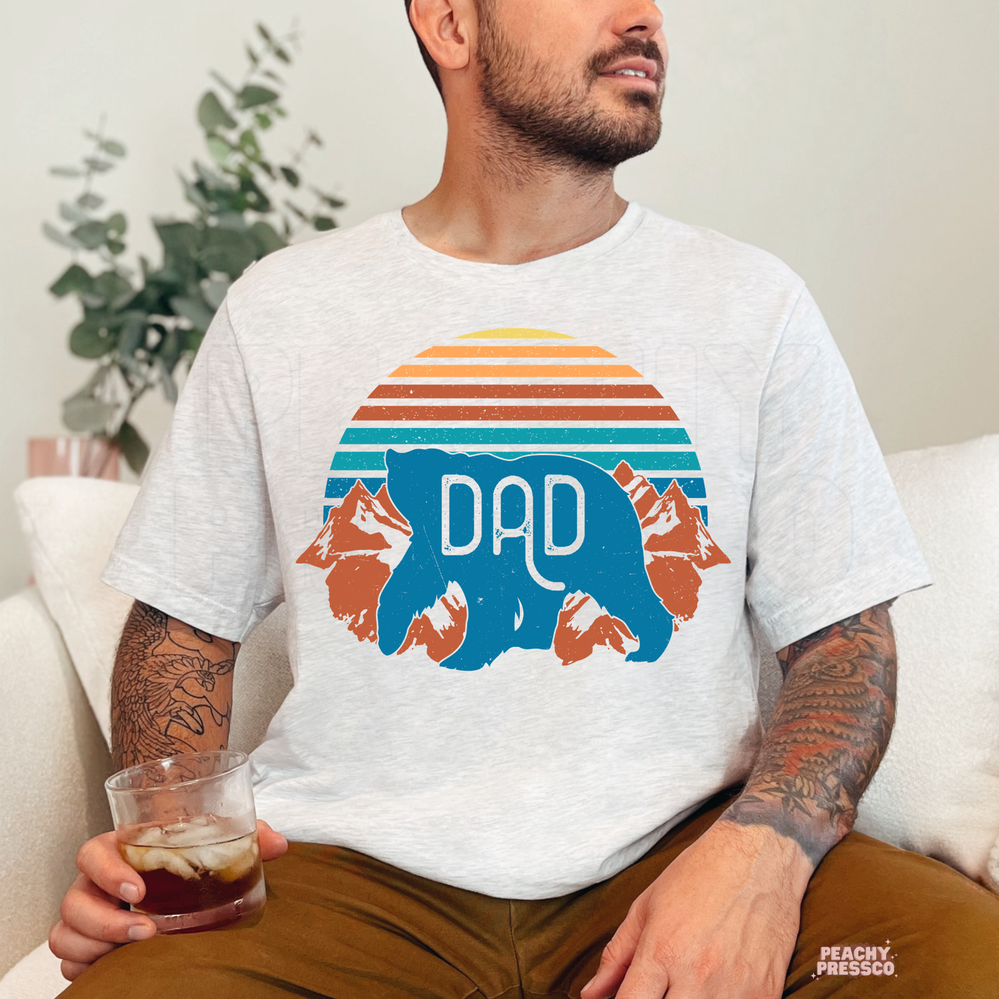 Man sitting indoors wearing ash grey t-shirt with vintage-style bear “Dad” graphic and retro sunset colors.