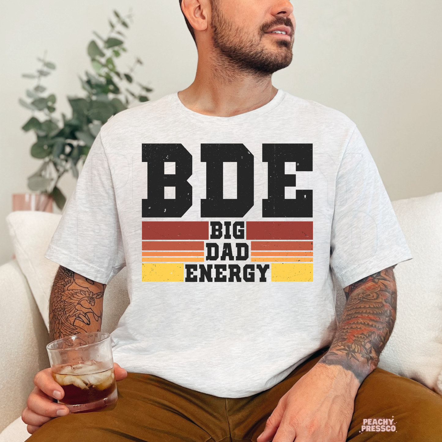 Man sitting indoors wearing ash grey t-shirt featuring bold “BDE Big Dad Energy” retro graphic print.