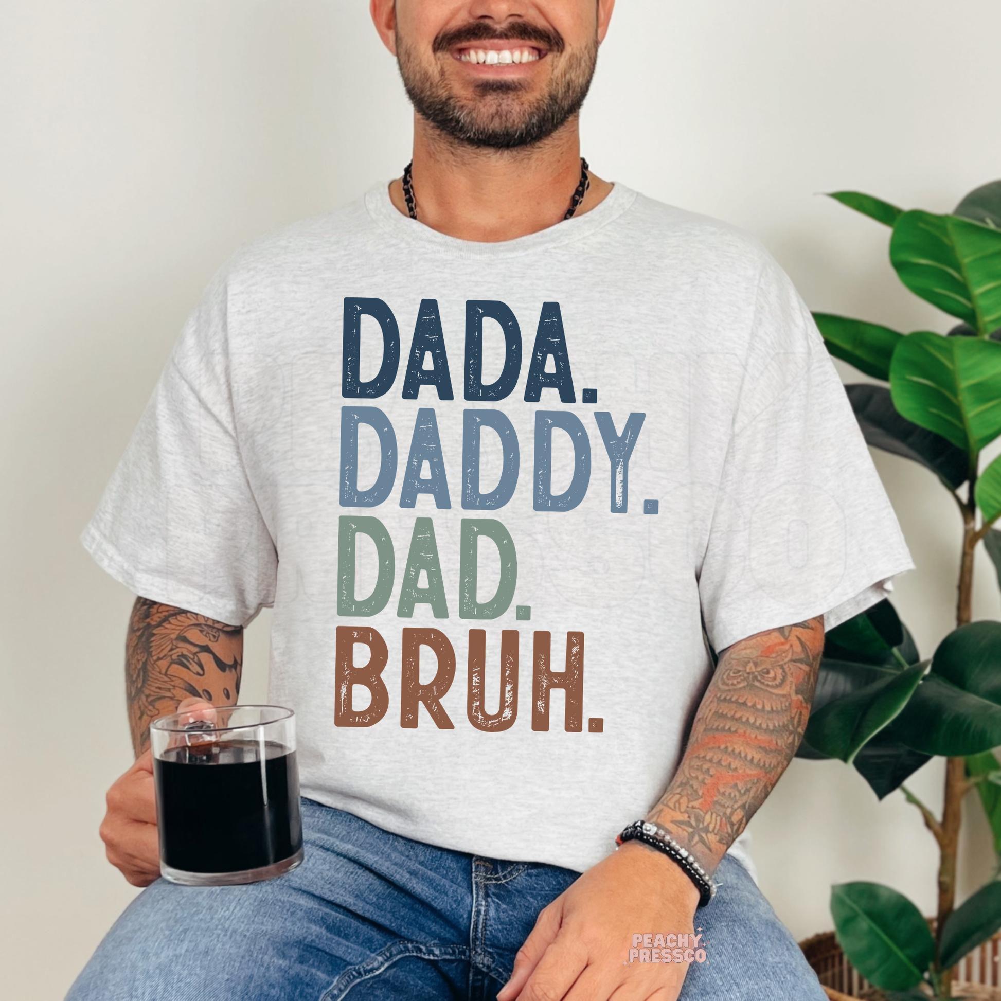 Man sitting indoors wearing ash grey t-shirt with “Dada Daddy Dad Bruh” funny fatherhood graphic design.