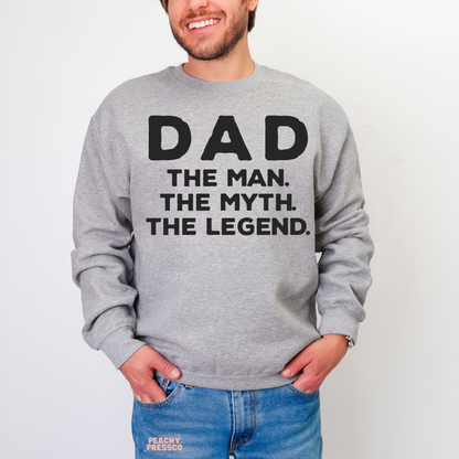 Man modeling grey crewneck sweatshirt featuring black “Dad The Man. The Myth. The Legend.” slogan design.