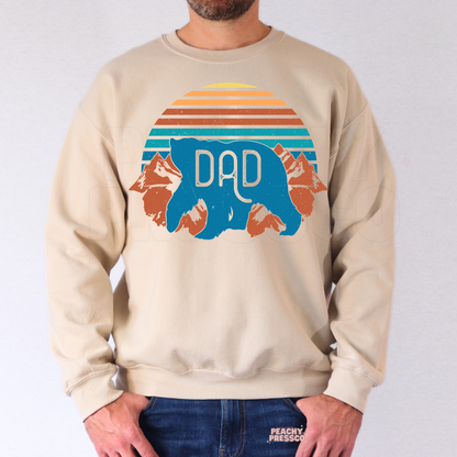 Man modeling beige crewneck sweatshirt with retro outdoors-inspired bear “Dad” graphic for Father’s Day gift.