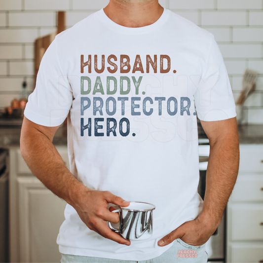 Man wearing white t-shirt with “Husband Daddy Protector Hero” colorful stacked typography graphic.