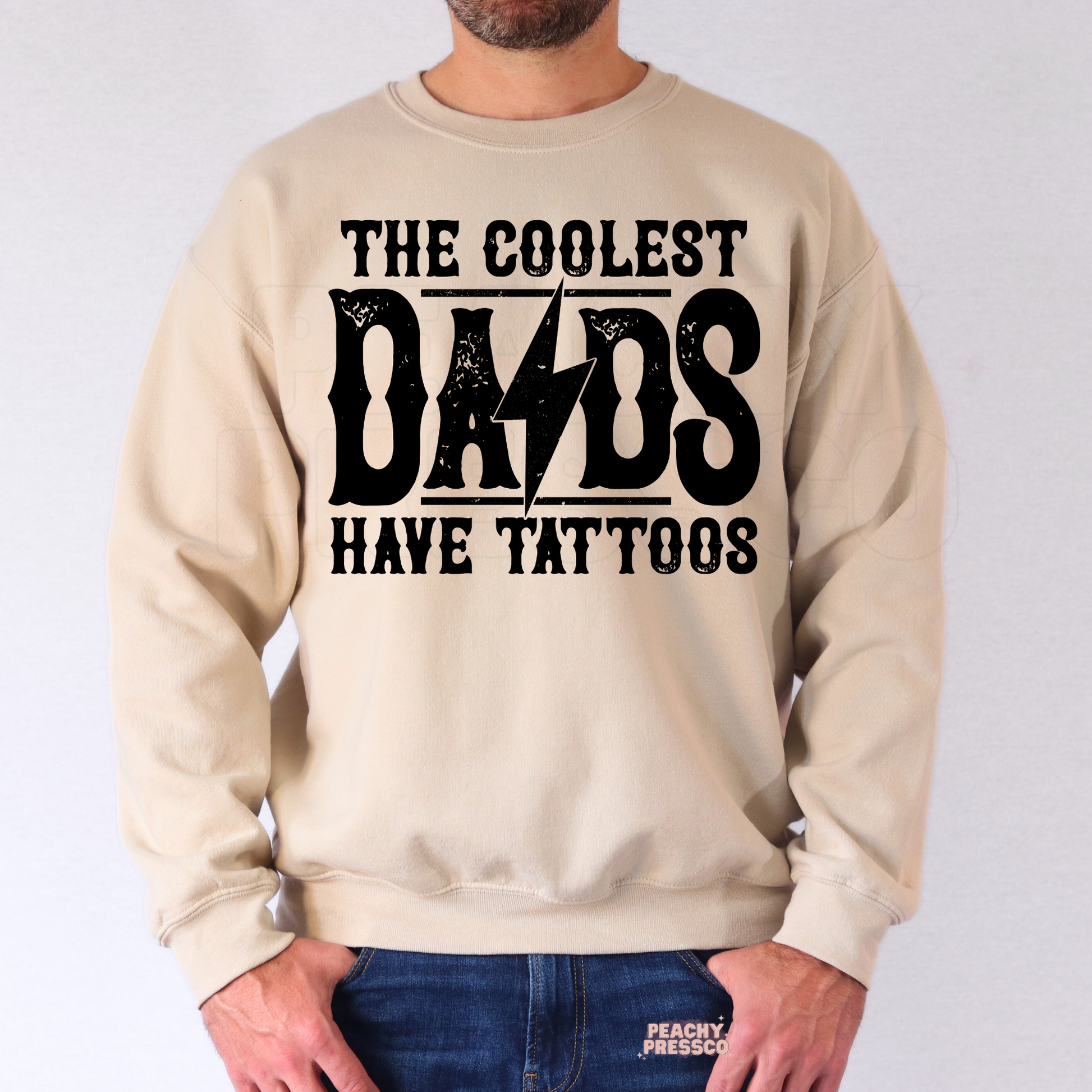 Man wearing beige crewneck sweatshirt with “The Coolest Dads Have Tattoos” graphic in bold vintage lettering.