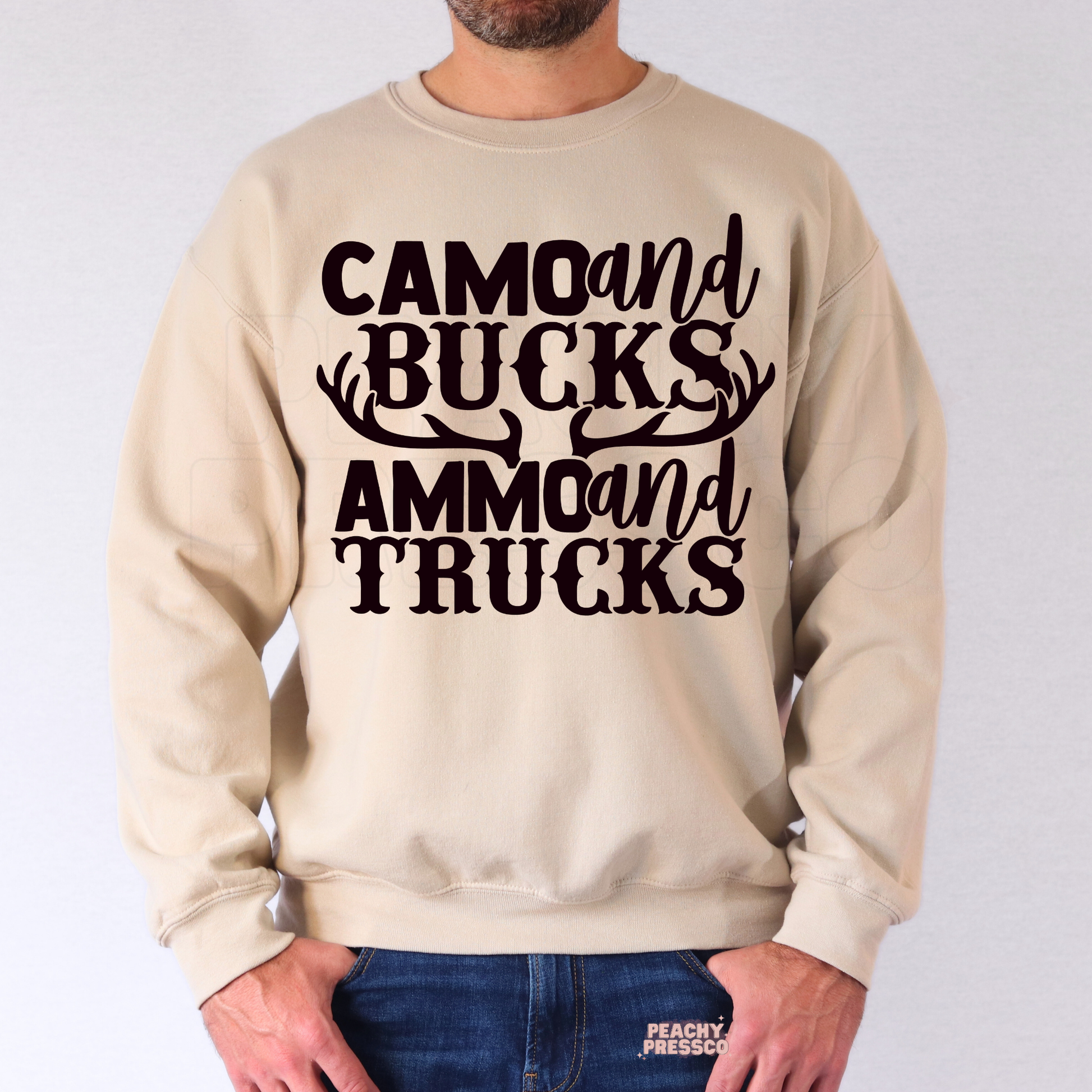 Man modeling beige crewneck sweatshirt with western hunting-inspired “Camo and Bucks Ammo and Trucks” text graphic.